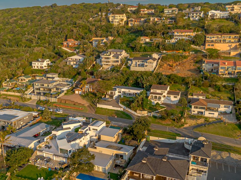 An aerial view of a coastal residential neighborhood featuring various houses nestled into a hillside with surrounding greenery and road infrastructure.