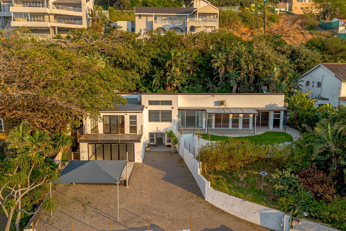 Aerial view of a multi-level residential property featuring a paved driveway, covered patio, balcony, and terraced garden areas.