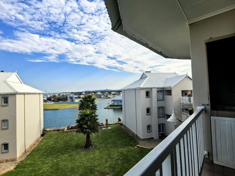 A covered balcony featuring tiled flooring and a metal railing, offering scenic views of the surrounding estate, canal, and landscaped garden area.