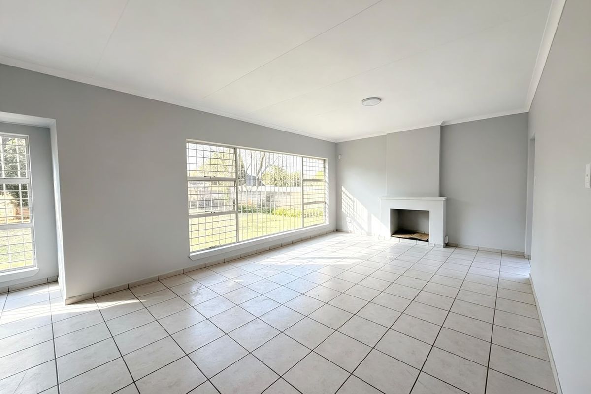 Spacious, light-filled living room featuring neutral tiled flooring, a built-in fireplace, and large windows with security bars overlooking the garden.