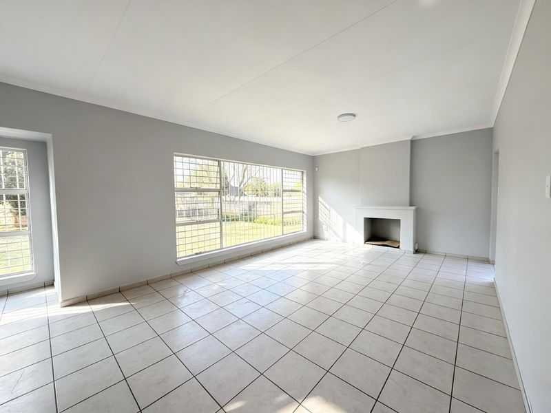 Spacious, light-filled living room featuring neutral tiled flooring, a built-in fireplace, and large windows with security bars overlooking the garden.