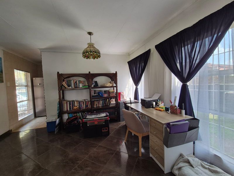 A functional home office space featuring tiled flooring, large windows with security bars, and natural light, suitable for a dedicated workspace.