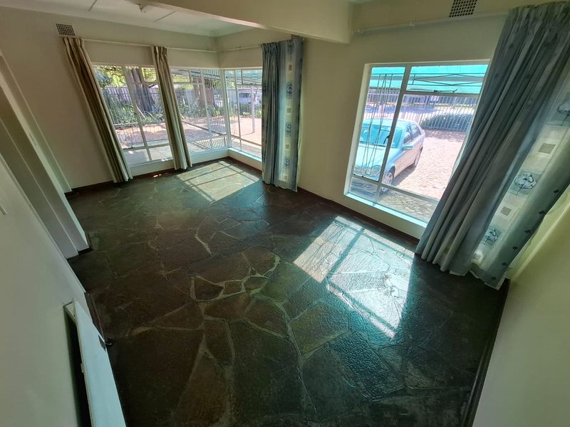 A bright, empty living room featuring natural flagstone flooring and large windows with security bars, offering a clear view of the exterior.