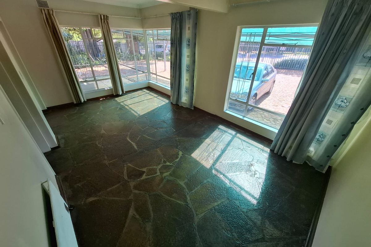A bright, empty living room featuring natural flagstone flooring and large windows with security bars, offering a clear view of the exterior.