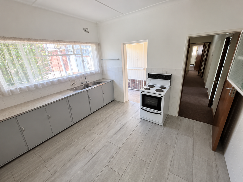 A functional kitchen featuring grey cabinetry, a double basin sink, and a freestanding electric stove, with neutral tiled flooring and direct access to a hallway.