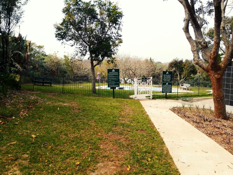 A fenced swimming pool area featuring a concrete pathway, mature trees, and safety signage within a residential estate setting.