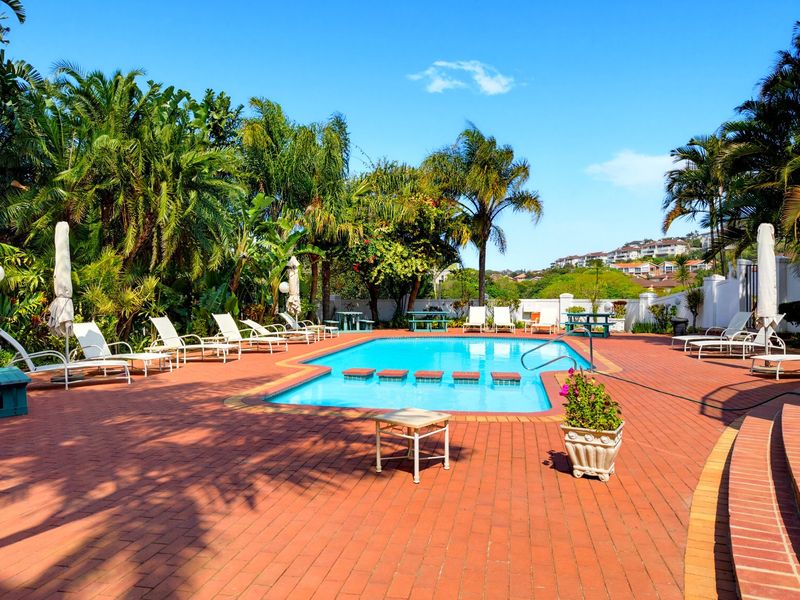 A well-maintained outdoor swimming pool area featuring brick paving, surrounding lush tropical landscaping, and a clear view of the neighborhood.