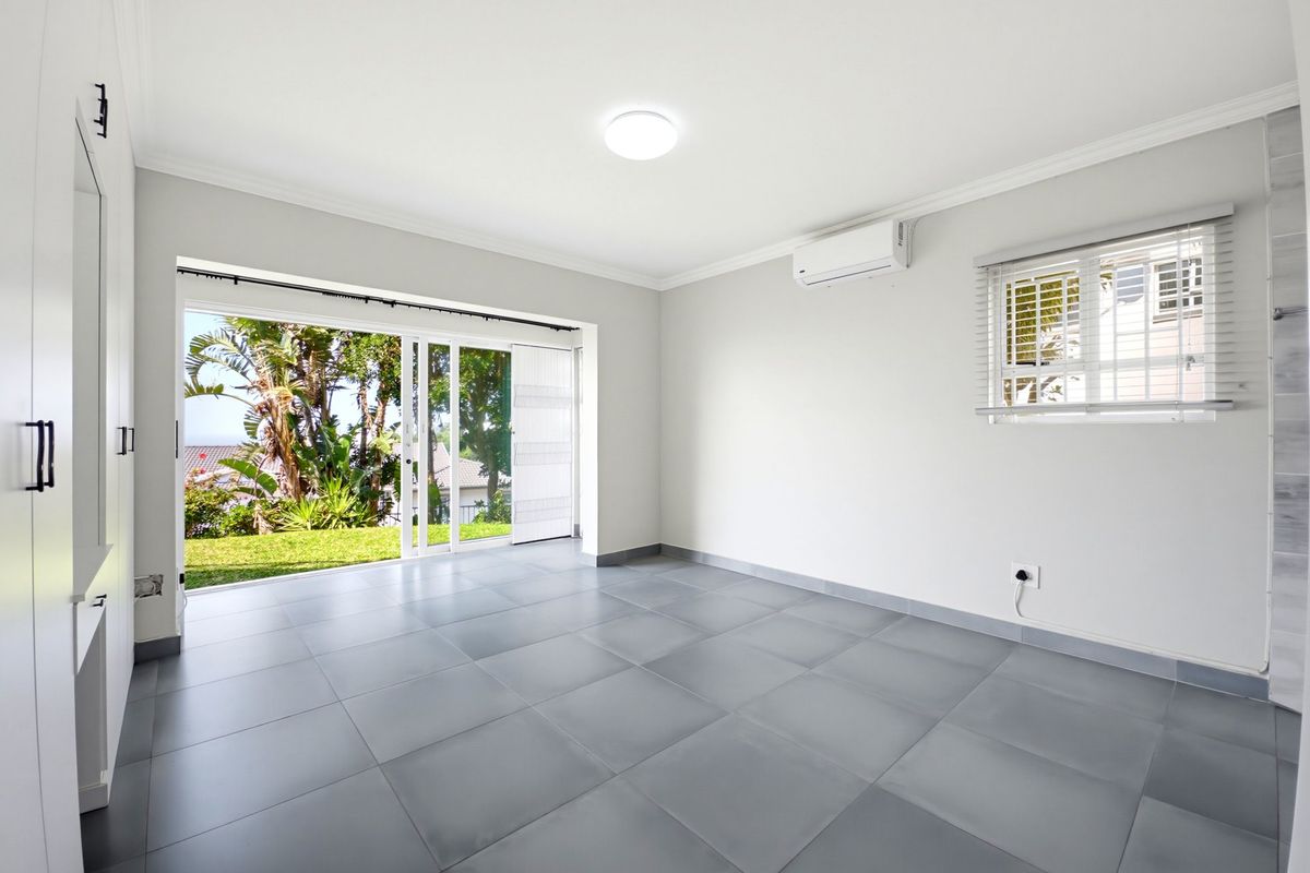 A bright, empty living room featuring large sliding glass doors opening to a garden, tiled flooring, and a wall-mounted air conditioning unit.