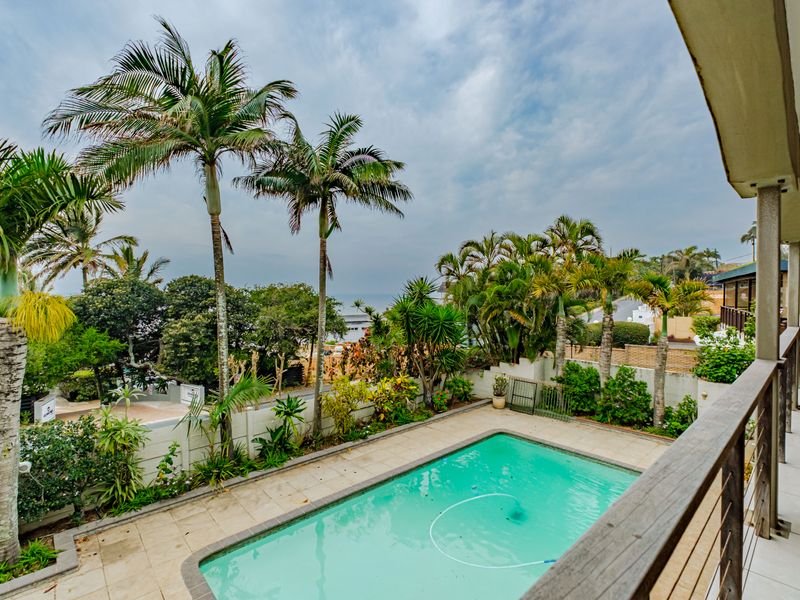 An elevated view of a swimming pool area with a paved deck, surrounded by lush palm trees and a boundary wall.