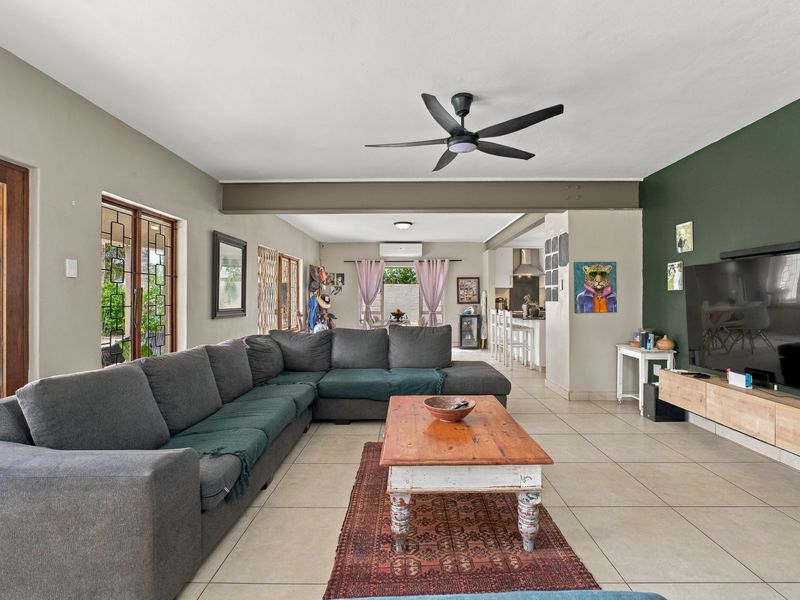 Spacious open-plan living area featuring tiled floors, a ceiling fan, exposed structural beam, and seamless flow to the kitchen and outdoor patio.