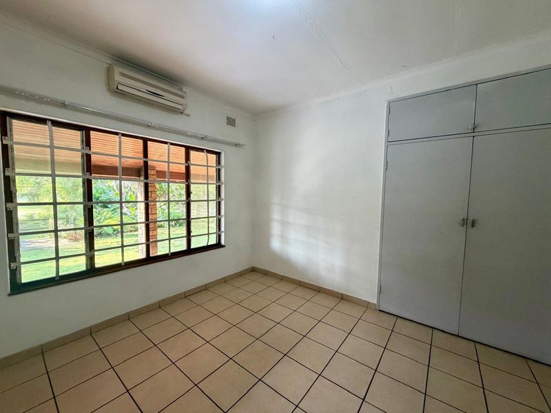 A bright, empty bedroom featuring tiled flooring, built-in cupboards, and an air conditioning unit, with security bars on the window.
