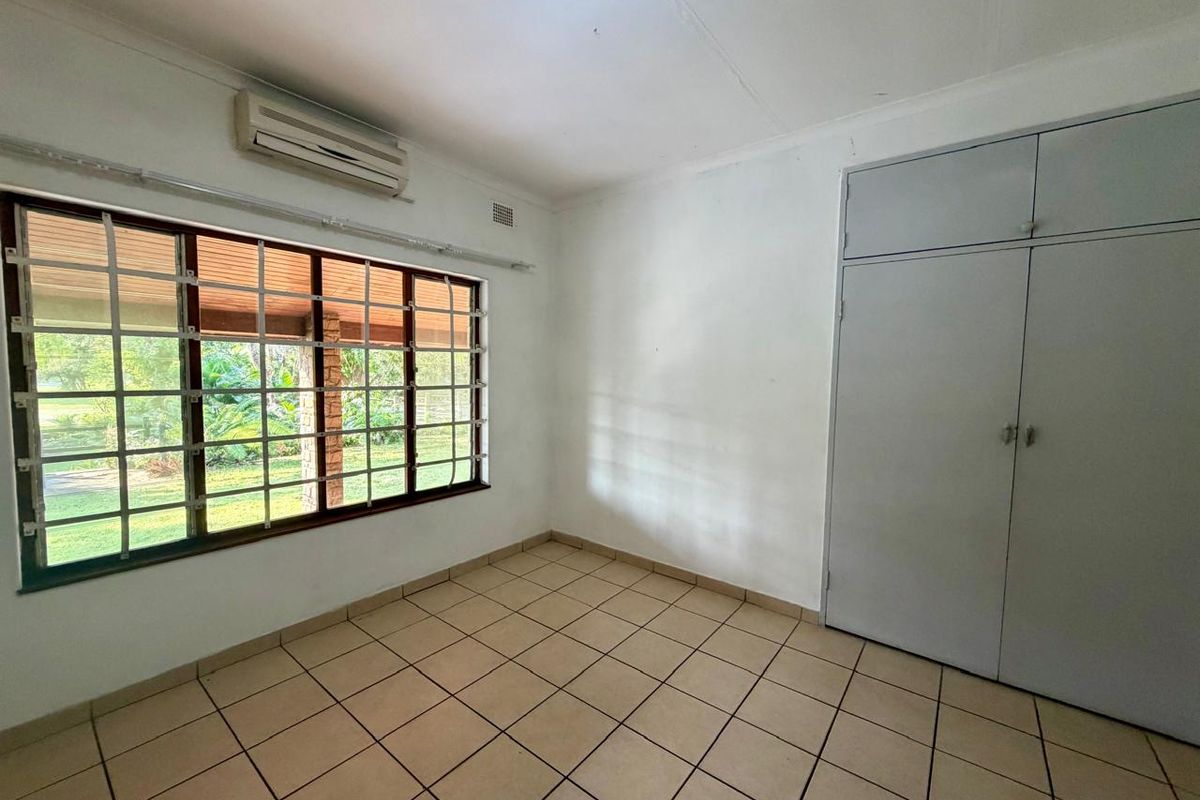 A bright, empty bedroom featuring tiled flooring, built-in cupboards, and an air conditioning unit, with security bars on the window.