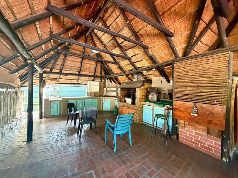 Covered outdoor patio featuring a traditional thatched roof, exposed timber rafters, brick flooring, and a built-in braai area with integrated cabinetry and sink.