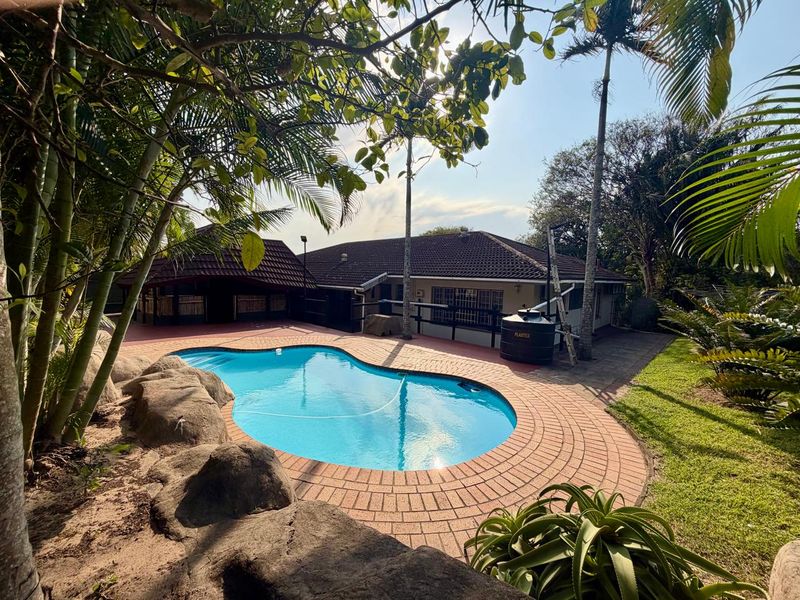 A well-maintained outdoor pool area featuring a paved surround, established garden landscaping, and a rear view of the main house structure.
