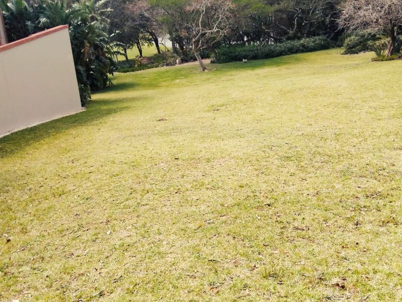A spacious, well-maintained residential garden area featuring a gentle slope, established trees, and a retaining wall.