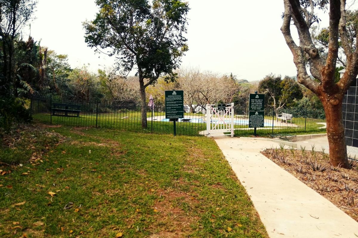 A fenced swimming pool area featuring a concrete pathway, mature trees, and safety signage within a residential estate setting.