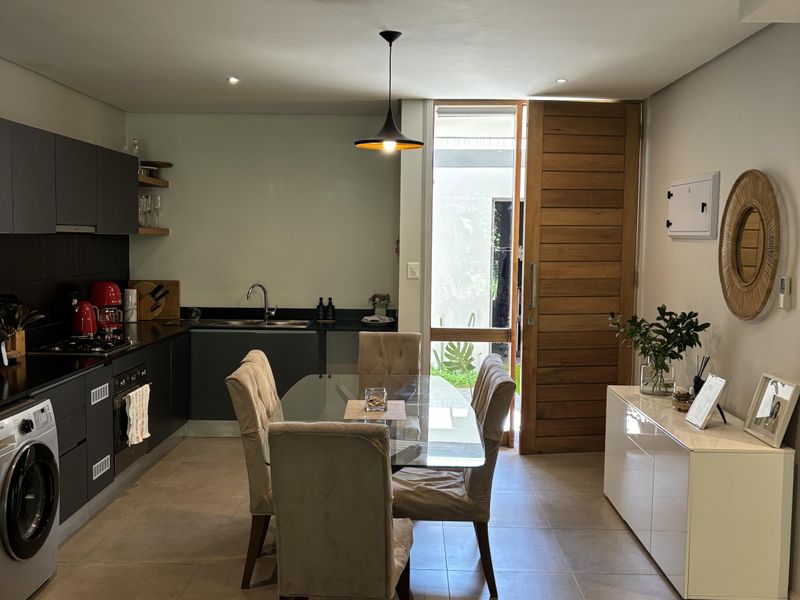 Modern open-plan kitchen and dining area featuring sleek dark cabinetry, tiled flooring, recessed lighting, and direct access to an outdoor space via a timber door.