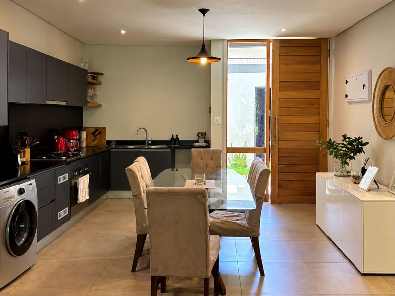 Modern open-plan kitchen and dining area featuring sleek dark cabinetry, integrated appliances, tiled floors, and direct access to an outdoor space via a timber door.