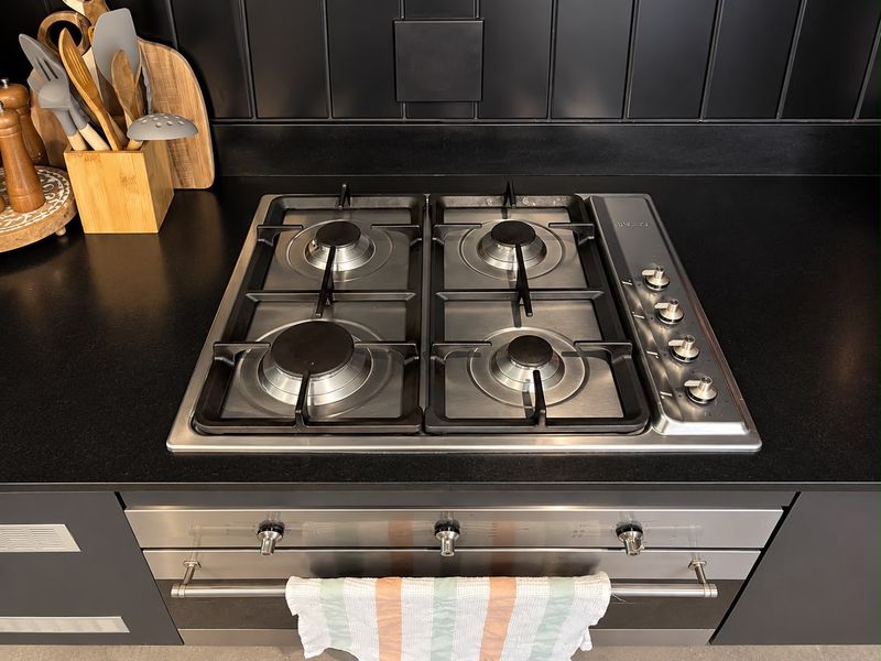 A modern kitchen featuring a stainless steel gas cooktop and built-in oven set against a sleek black countertop and tiled splashback.