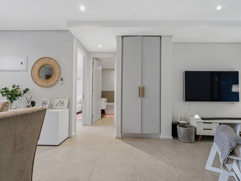 A well-maintained living area featuring neutral tiled flooring, recessed lighting, and a built-in storage cupboard, with a clear view into the adjacent hallway and bathroom.