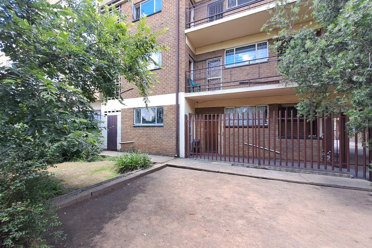 Rear view of a multi-story brick apartment building featuring balconies, security fencing, and a paved courtyard area.