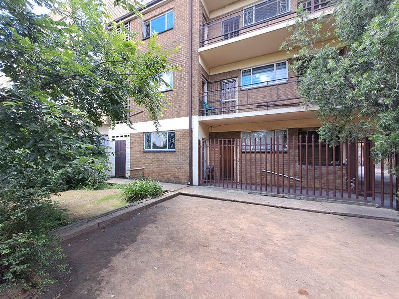 Rear view of a multi-story brick apartment building featuring balconies, security fencing, and a paved courtyard area.