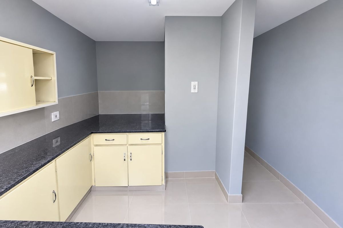 A clean, empty kitchen area featuring granite countertops, light-colored built-in cabinetry, and neutral tiled flooring with a modern grey wall finish.
