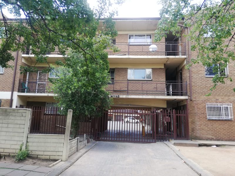 Front elevation of a multi-story brick apartment building featuring gated driveway access, balconies with metal railings, and security bars on windows.