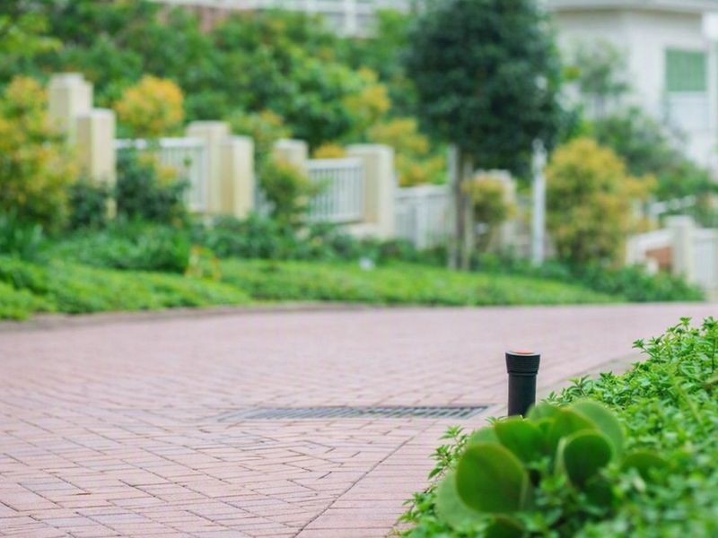 A view of a paved residential driveway featuring a built-in stormwater drain and irrigation sprinkler system, bordered by landscaped garden beds and boundary walls.