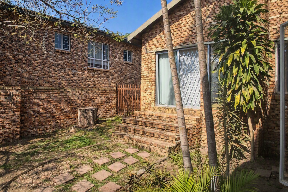 Rear exterior view of a face brick house featuring a small garden, paved walkway, and brick steps leading to a sliding glass door.