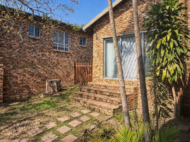 Rear exterior view of a face brick house featuring a small garden, paved walkway, and brick steps leading to a sliding glass door.