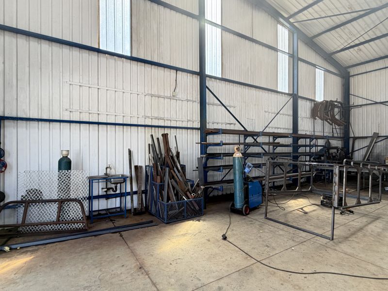 A functional industrial utility space or workshop featuring a steel portal frame, corrugated metal walls, concrete flooring, and integrated metal storage racking.