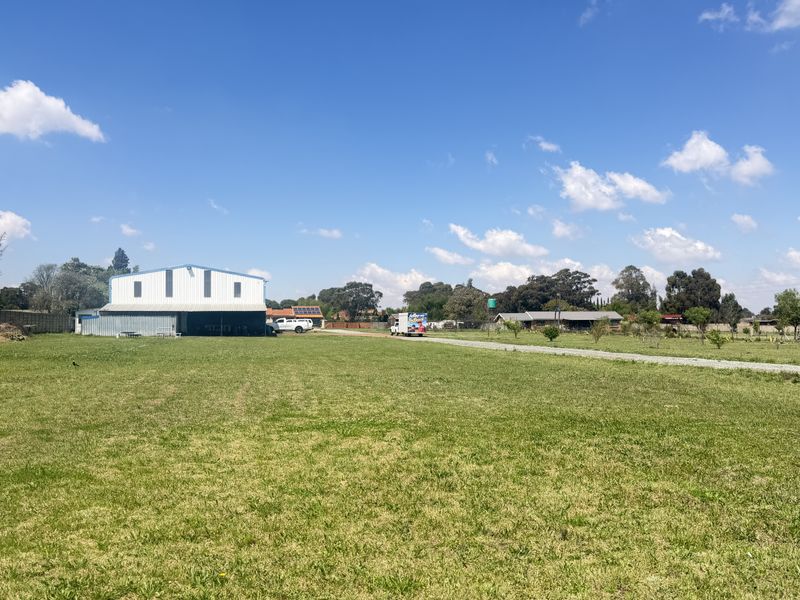 A wide-angle view of the small holding property featuring a large metal shed, expansive open grass areas, and a gravel driveway.