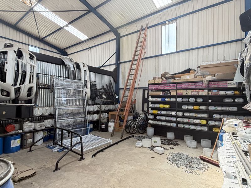 A large industrial utility space featuring a steel portal frame, corrugated metal walls, skylights, and extensive built-in shelving for storage.