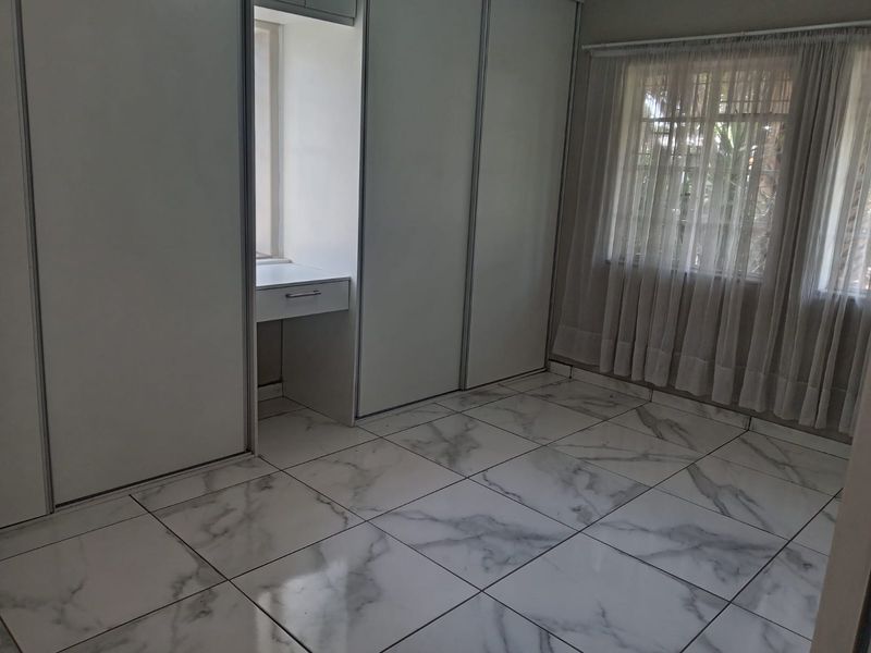 A bright, empty bedroom featuring modern marble-look floor tiles, extensive built-in cupboards, and a convenient integrated vanity area.