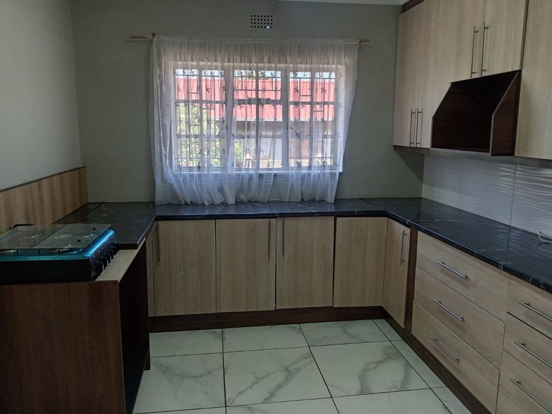 A clean, functional kitchen featuring light wood-finish cabinetry, dark countertops, tiled flooring, and a window with security bars.