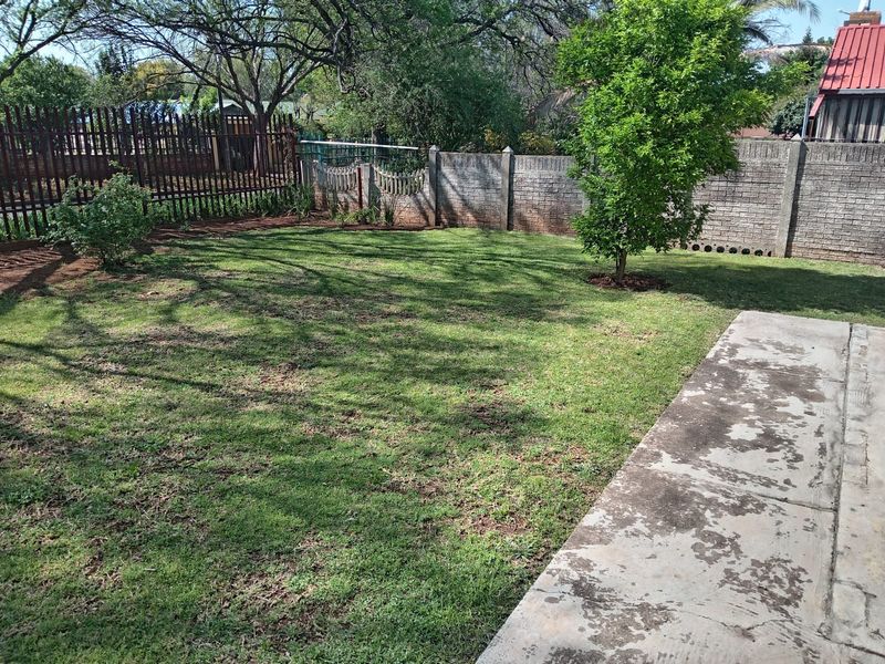 A private backyard featuring a grass lawn, mature trees, a concrete patio area, and secure perimeter walling with metal fencing.