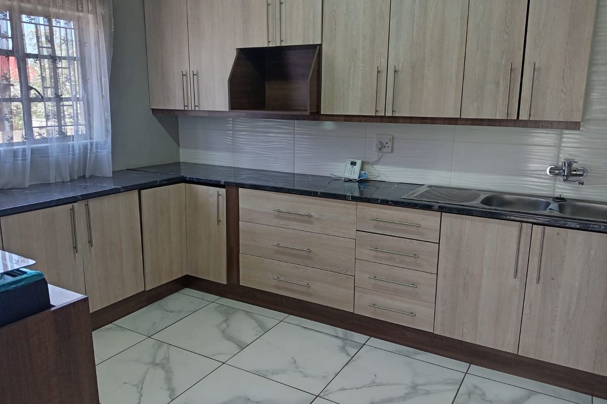 A clean, modern kitchen featuring light wood-grain cabinetry, dark stone-look countertops, a stainless steel sink, and white marble-effect floor tiles.