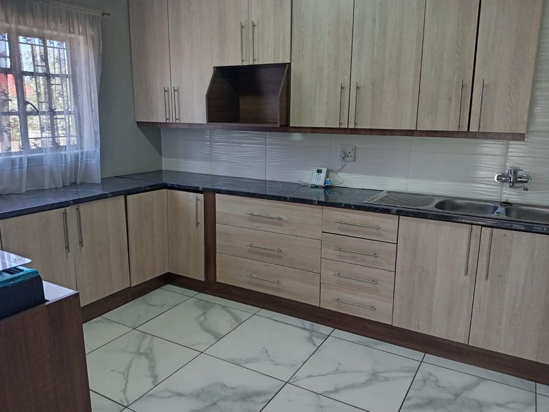 A clean, modern kitchen featuring light wood-grain cabinetry, dark stone-look countertops, a stainless steel sink, and white marble-effect floor tiles.
