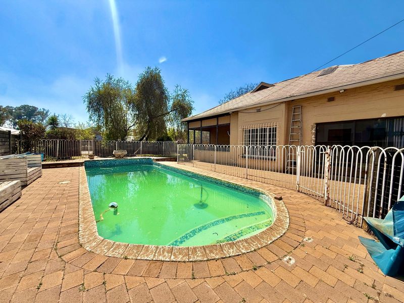 Outdoor pool area featuring a paved deck, metal security fencing, and a swimming pool requiring maintenance and cleaning.