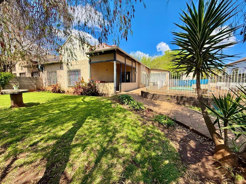 Rear view of the property featuring a covered patio, paved walkway, and a swimming pool set within a landscaped garden.