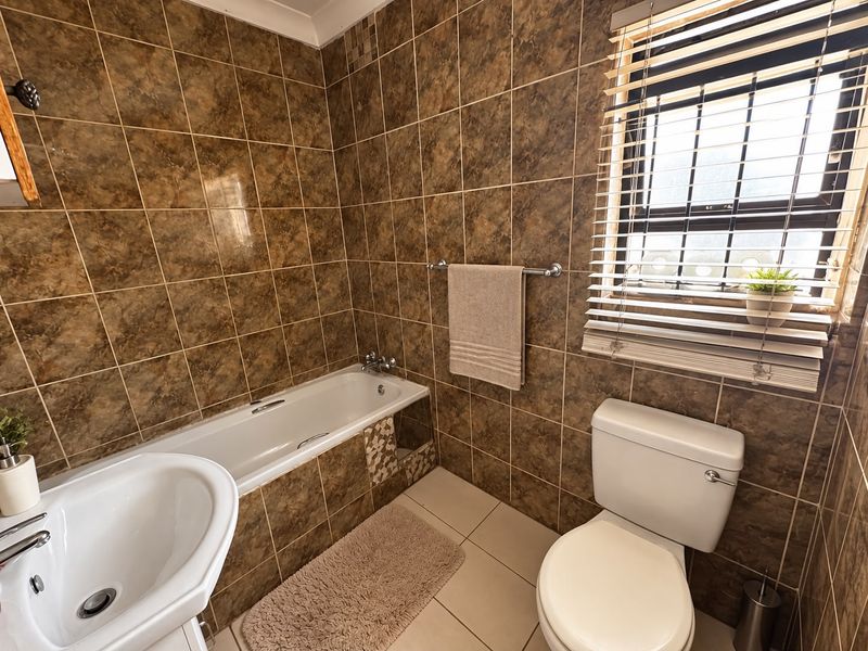A functional bathroom featuring full-height brown marble-effect wall tiles, a standard bathtub, pedestal basin, and toilet, complemented by natural light from a window with blinds.