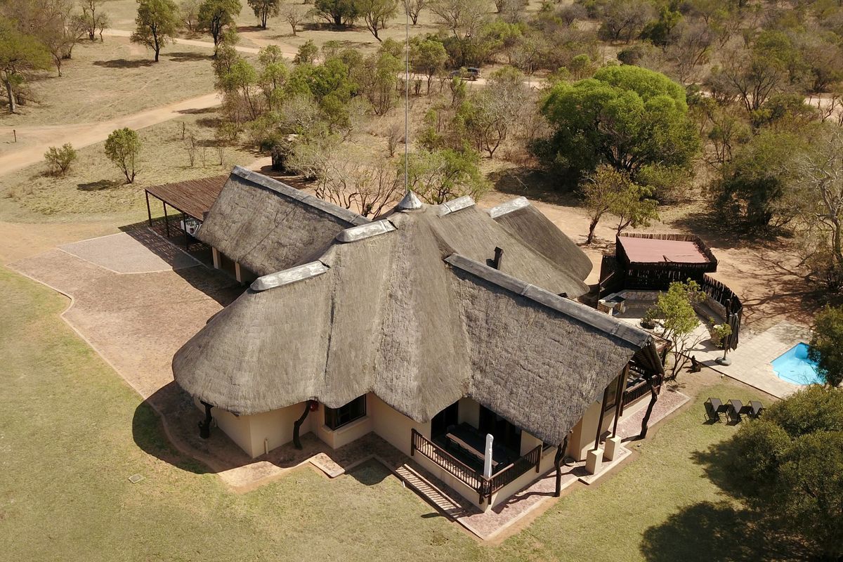 Aerial view of a lodge property featuring a traditional thatch roof, swimming pool, and surrounding natural bushveld landscape.