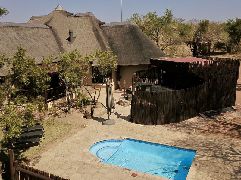 Aerial view of a lodge exterior featuring a swimming pool, paved deck, boma area with timber fencing, and a traditional thatched roof structure.
