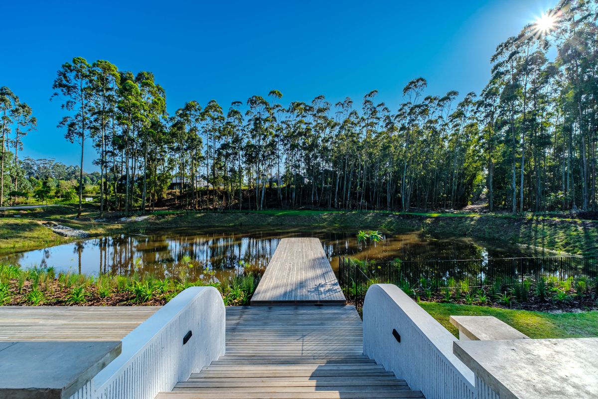 A beautifully landscaped rear garden featuring a timber deck walkway leading to a serene pond, surrounded by mature trees and modern concrete retaining walls.