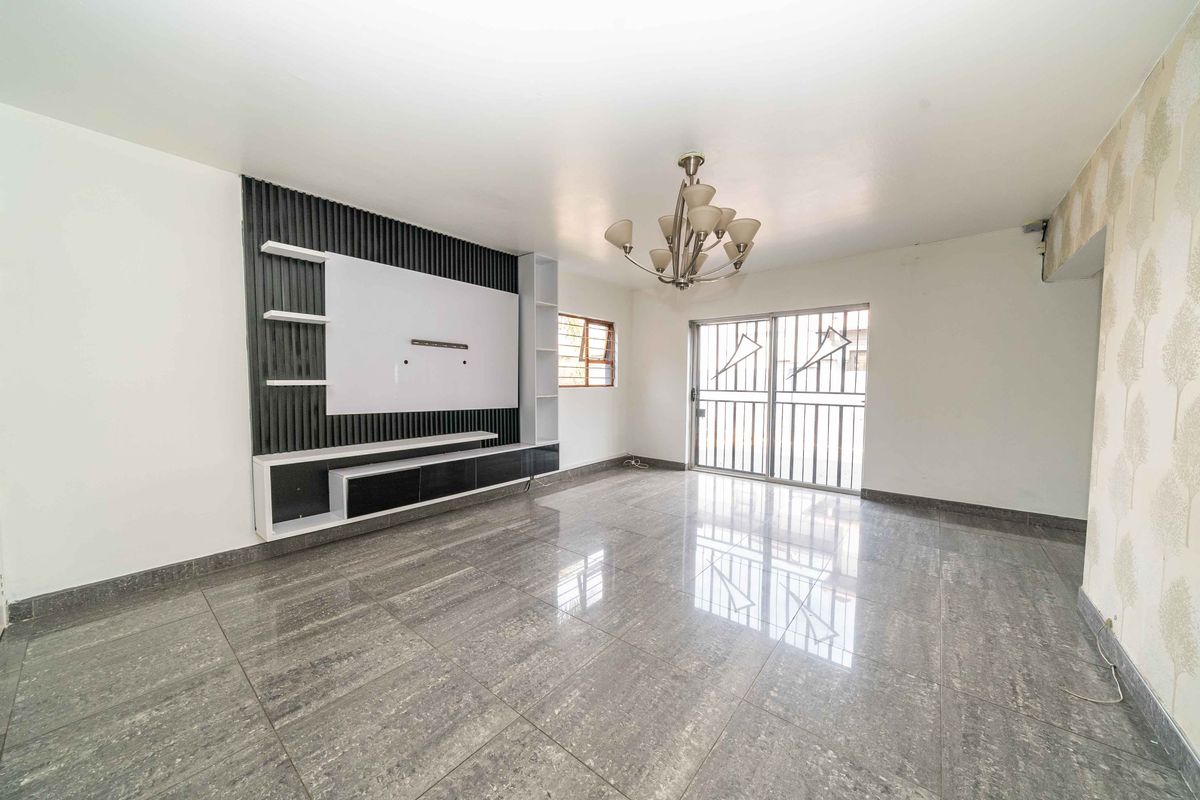 Spacious living room featuring polished tiled flooring, a modern built-in media unit, and a decorative chandelier, with access to an outdoor area via sliding doors.