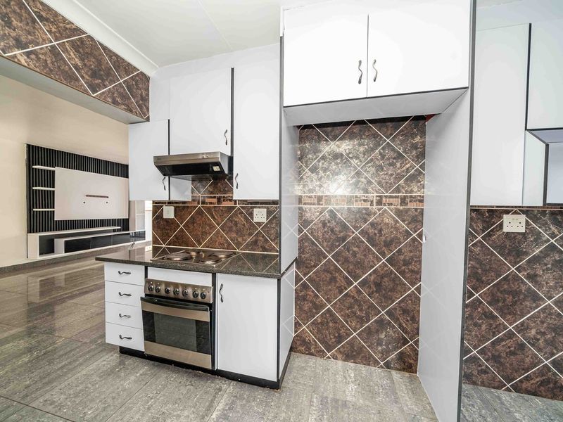 A clean, empty kitchen featuring white cabinetry, a built-in oven with hob, extractor fan, and dark patterned tiled backsplash and flooring.