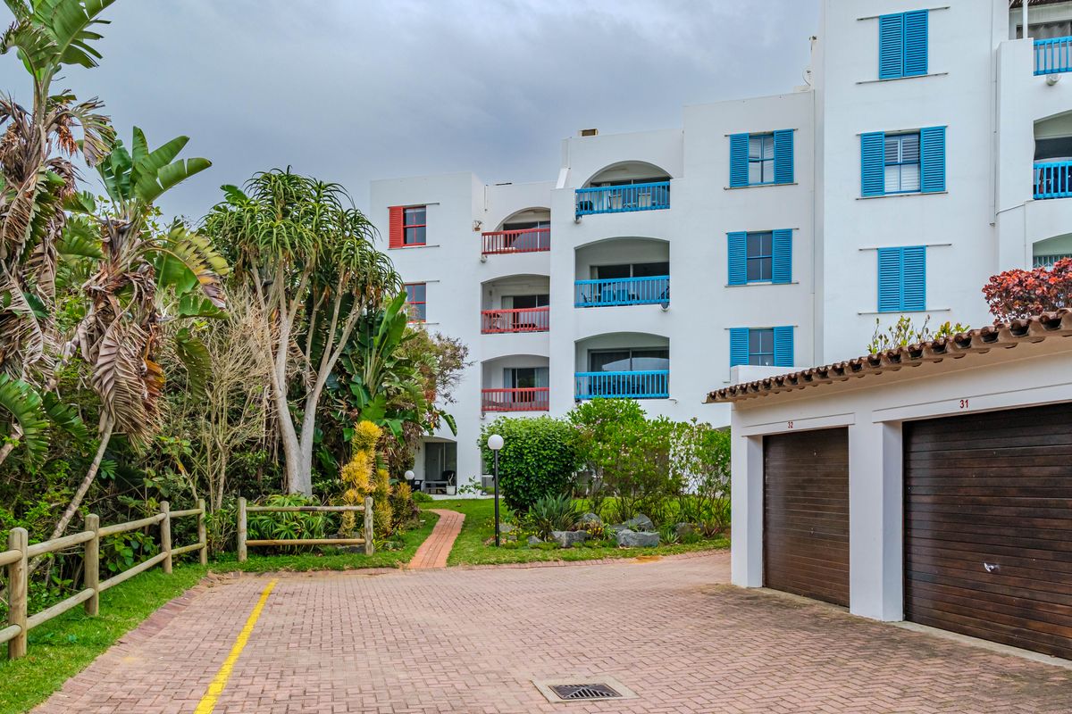Exterior view of an apartment complex featuring multiple balconies with colorful shutters, a paved driveway, and private garage units set within a landscaped garden.