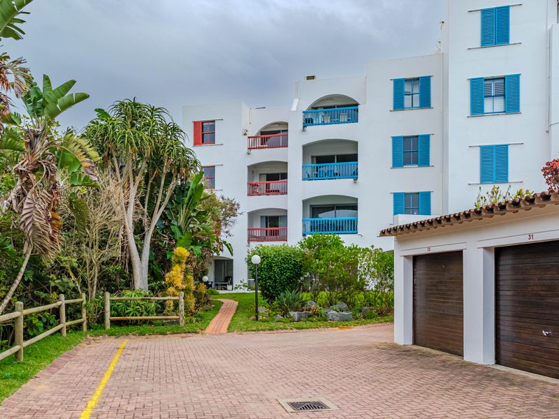 Exterior view of an apartment complex featuring multiple balconies with colorful shutters, a paved driveway, and private garage units set within a landscaped garden.