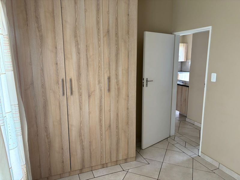 A bedroom featuring light wood-grain built-in wardrobes and tiled flooring, with a doorway leading to an adjacent kitchen area.